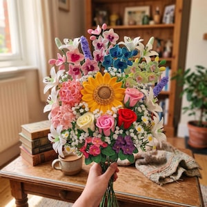 Könnte beinhalten: Ein lebendiger Strauß gehäkelter Blumen, darunter eine große gelbe Sonnenblume, rosa Rosen, weiße Lilien und verschiedene andere bunte Blüten. Der Strauß wird in einer Hand gehalten und zeigt die detaillierte Handwerkskunst.