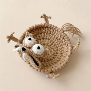 May include: A tan crocheted coaster in the shape of a cartoon character with large white eyes, a wide mouth, and small limbs. The coaster has a textured appearance and a tail made of loose yarn.