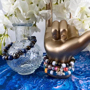 May include: A clear glass vase holds a beaded bracelet with blue and brown beads. A gold hand sculpture displays several beaded bracelets in various colors. White flowers are in the background. The bracelets are on a blue and white patterned surface.