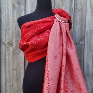 May include: A red baby carrier wrap with a star pattern, draped over a black mannequin. The wrap is secured with a silver ring. The fabric has a lighter red border and is decorated with small red stars.
