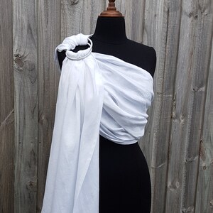 May include: White fabric baby wrap with a silver ring on a black mannequin. The wrap is draped over the mannequin's shoulder and tied around the waist.