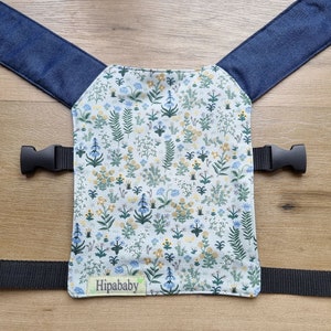 May include: A baby carrier with denim straps and a floral fabric panel. The panel features a white background with a blue and yellow floral pattern. The carrier has a black strap with a buckle and the text "Hipababy" is printed on a green tag.