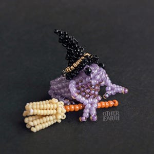 Könnte beinhalten: Eine Perlen-Hexenfrosch-Figur mit schwarzem Hut und orangefarbenem Besen. Der Frosch ist lila mit goldenen Akzenten. Der Besen ist beige und orange. Der Text "OTHER EARTH" befindet sich auf dem Besen.