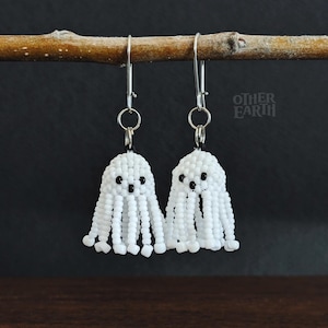 May include: Two white beaded ghost earrings with black bead eyes and dangling beaded strands. The earrings are suspended from silver-coloured ear wires and hang from a small branch. The background is dark, and the earrings are the focal point.