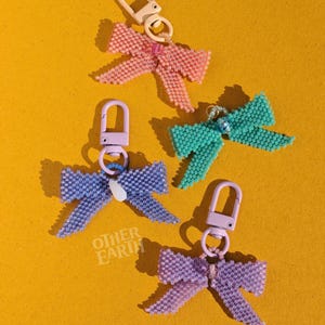 May include: Four beaded bow keyrings in pink, teal, purple, and blue, each with a metal clasp. The keyrings are arranged on a yellow background. The text "OTHER EARTH" is visible on one of the bows.