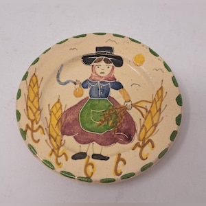 May include: Hand-painted ceramic plate depicting a woman in traditional dress, holding wheat and a small bag. The plate has a cream background with green and yellow accents, including wheat stalks and a dotted border. A sun and birds are also shown.