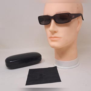 May include: Dark brown rectangular sunglasses with tinted lenses are displayed on a mannequin head. A black case and cleaning cloth are next to the sunglasses. The sunglasses have a subtle logo on the temple.