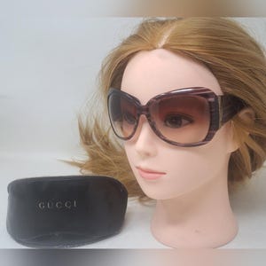 May include: Oversized, square-framed sunglasses with a gradient brown tint. The frames are a dark purple with a striped pattern and gold accents. A black case with the word "GUCCI" is also visible.