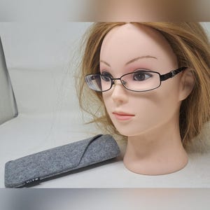 May include: Black-framed eyeglasses with rectangular lenses are displayed on a mannequin head. A gray felt eyeglass case is next to the mannequin. The case has the word "KITO" printed on it.