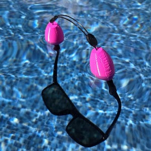 Op de afbeelding: Zonnebril met zwart montuur en donkere glazen is verbonden met twee roze drijvers door een zwart koord. De drijvers bevinden zich in het water en de zonnebril drijft op het oppervlak van een zwembad.