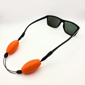 Puede incluir: Gafas de sol negras con lentes oscuras y una correa negra. La correa tiene dos flotadores oblongos de color naranja brillante con el texto "FLOATROP". Sobre un fondo blanco.