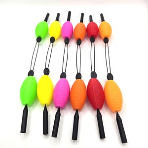 Könnte beinhalten: Sechs Angelposen in einer Reihe angeordnet, jede mit einem doppelten ovalen Design, das durch eine schwarze Schnur verbunden ist. Die Posen sind in Neongrün, Gelb, Pink, Orange, Rot und Hellorange gehalten, mit schwarzen Kunststoffspitzen.