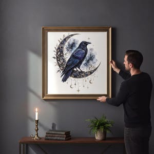 A man is hanging a framed artwork of a black crow on a wall, with a lit candle and some books on a table below it.