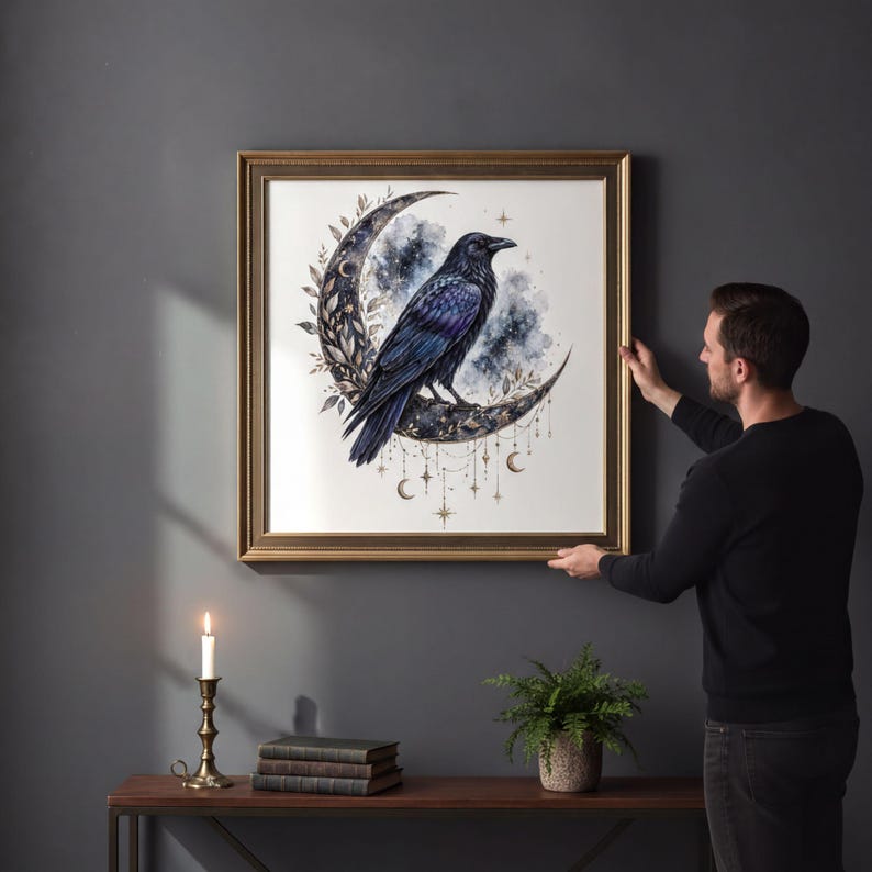 A man is hanging a framed artwork of a black crow on a wall, with a lit candle and some books on a table below it.