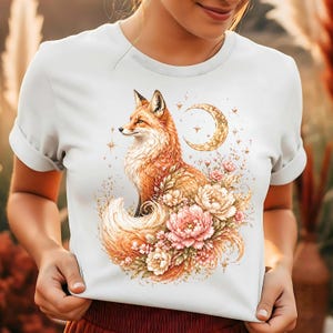 May include: A white short-sleeved t-shirt featuring a detailed illustration of a red fox sitting among soft pink and cream-colored flowers. Above the fox is a golden crescent moon surrounded by small sparkling stars, creating a celestial design.