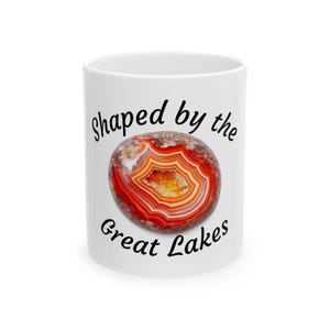May include: White ceramic mug featuring a colorful agate stone design with red, orange, and yellow hues. The mug has the text "Shaped by the Great Lakes" in black script, making it a unique gift or souvenir.