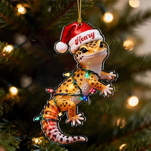 Peut inclure: Ornement de Noël représentant un gecko souriant portant un bonnet de Père Noël avec le nom "Henry" et enroulé de guirlandes lumineuses colorées. Le gecko a un motif tacheté jaune et orange. L'ornement est suspendu à une ficelle dorée sur un fond de sapin de Noël flou.