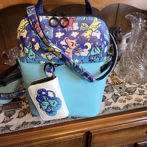May include: A light blue handbag with a dragon-themed design. The top section features a pattern of dragons, stars, and moons on a dark blue background. Includes a matching strap and a small white accessory with a dragon graphic.