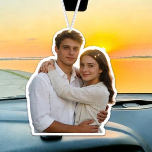May include: Car air freshener featuring a couple embracing, set against a sunset over a body of water. The photo is printed on a white, die-cut, car accessory, hanging from a white string. The dashboard and windshield are visible.
