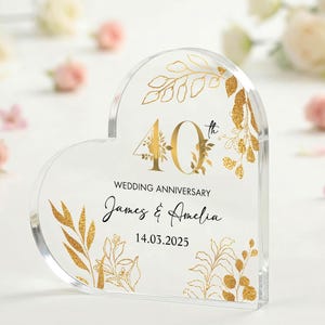May include: Clear acrylic heart-shaped wedding anniversary keepsake. Features gold floral accents and the text "40th Wedding Anniversary" with names and a date. Surrounded by white and pink flowers.