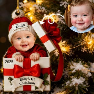 Puede incluir: Adorno navideño con un bebé con un suéter rojo y un gorro de Papá Noel con el nombre "Dana". El bebé está dentro de una caja de regalo con un lazo rojo y el texto "Baby's 1st Christmas" y el año 2025. Una foto del bebé está en el fondo.