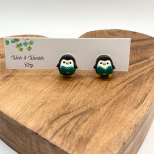 May include: A pair of penguin-shaped stud earrings. Each earring features a black and white penguin with a green scarf and yellow feet. The earrings are displayed on a white card with the text "Wink & Wander Clay".