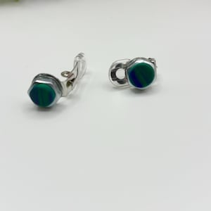 Puede incluir: Un par de pendientes de clip plateados. Cada pendiente presenta una piedra hexagonal verde y azul engastada en un marco de metal plateado. Los pendientes se muestran sobre una superficie blanca.