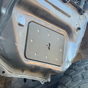 May include: A black rectangular panel with six silver screws is mounted on a gray metal surface. The panel has a keyhole-shaped cutout in the center. The image shows part of a vehicle's body and a tire.