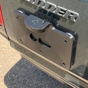 Puede incluir: Soporte de matrícula de metal negro fijado a un vehículo verde oscuro. El soporte tiene un diseño articulado con un asa y varios orificios de montaje. La palabra "4Runner" es visible en letras plateadas encima del soporte.