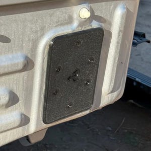 May include: A close-up of a section of a vehicle's exterior, showing a textured black rectangular plate secured with multiple screws. The plate is mounted on a metallic gray surface.