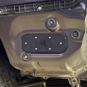 May include: A black rectangular panel with the letter "R" in white is attached to a vehicle's frame with silver screws. The panel is set against a gray and brown metallic surface, with the Toyota logo visible.