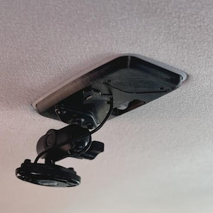 May include: A black, mounted camera system, likely for a vehicle, attached to a light-colored ceiling. The camera is adjustable and connected to a wire. The mount is rectangular with rounded edges.