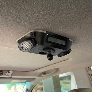 May include: A black car accessory mounted on a vehicle's ceiling. It features a rectangular display, a small LED light, and a ball mount. The accessory is designed to be installed in a car.