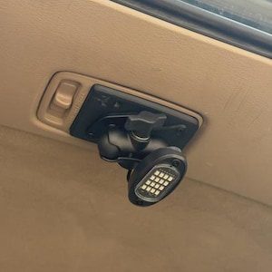 Può includere: Luce LED nera con una staffa di montaggio fissata a una superficie beige. La luce ha una griglia di piccole luci bianche. Sono visibili un braccio e una manopola neri regolabili. La staffa ha un interruttore.