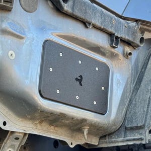 May include: A black metal plate with a stylized logo is mounted on a vehicle. The plate is rectangular with rounded corners and is secured with multiple silver bolts. The vehicle's body is a light gray color, and the plate is mounted near the tire.