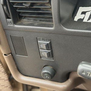 Op de afbeelding: Close-up van het dashboard van een voertuig met een zwart ventilatierooster, twee tuimelschakelaars en een zwarte knop. Het dashboard is donkerbruin. Een wit en zwarte sticker met de letters "FI" is zichtbaar.