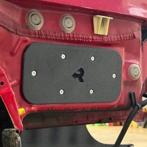 May include: A black rectangular panel with the letter "R" cut out, mounted on a red vehicle component. The panel is secured with silver screws. The vehicle's red body is visible, along with other components.