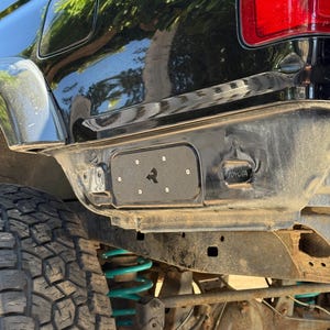 May include: Close-up of the rear of a black Toyota SUV. The image shows the bumper, taillight, and a portion of the tire. The suspension features teal springs. A black metal plate with a logo is mounted on the bumper.