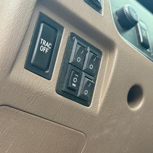 May include: Close-up of a vehicle's dashboard featuring a black "TRAC OFF" button and a cluster of four black rocker switches. The switches have white markings and are set against a tan interior.