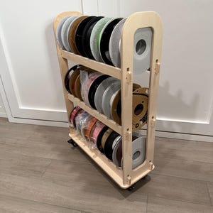 Może przedstawiać: Drewniany stojak do przechowywania filamentu do drukarki 3D na kółkach, mieszczący wiele szpul w różnych kolorach. Stojak ma trzy półki i naturalne drewniane wykończenie. Szpule są w odcieniach czerni, bieli, zieleni, brązu i różu.