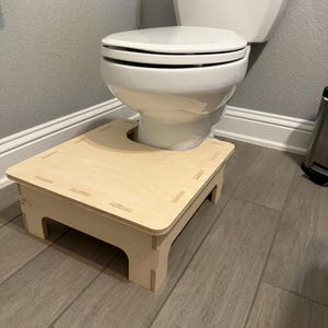 Puede incluir: Un taburete de inodoro de madera de color claro con un recorte para la base del inodoro. El taburete es rectangular con esquinas redondeadas y cuatro patas. Está colocado frente a un inodoro blanco.