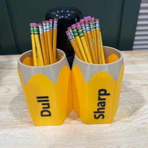 Op de afbeelding: Twee gele potloden in de vorm van een potlood met de tekst "Dull" en "Sharp" bevatten potloden met roze gummen. De containers hebben een bruine bovenkant en staan op een houten oppervlak. Een zwarte vaas staat op de achtergrond.
