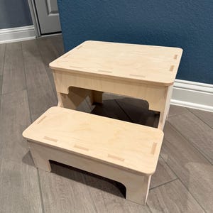 Puede incluir: Un taburete de madera de dos escalones con acabado natural. El taburete tiene una parte superior rectangular y un escalón inferior, ambos con veta de madera clara. El diseño presenta asas recortadas y es adecuado para diversos usos.