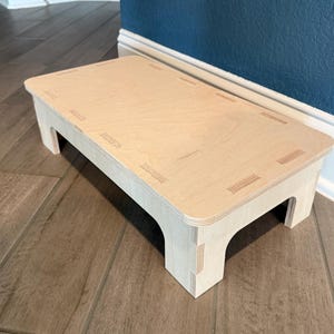 May include: A light-colored wooden step stool with a rectangular top and four sturdy legs. The step stool is made of light-colored wood and has a simple, functional design. The step stool measures approximately 18 inches long, 12 inches wide, and 8 inches high.
