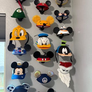 Op de afbeelding: Een verzameling hoeden met personagethema's, waaronder Mickey Mouse en Donald Duck, wordt tentoongesteld op een grijze muur. De hoeden hebben iconische ontwerpen en kleuren, met bevestigde zwarte oren. Sommige hoeden hebben extra karakterdetails.