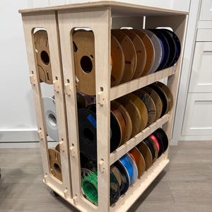 Puede incluir: Un carrito de almacenamiento de madera clara con múltiples estantes verticales y horizontales diseñados para sostener bobinas circulares. La unidad presenta una construcción modular con juntas visibles y está montada sobre ruedas negras.