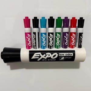 May include: A set of eight Expo dry erase markers in various colors including pink, black, blue, green, purple, and red, arranged in a horizontal white plastic holder. The holder features the black and white Expo low odor chisel tip logo.