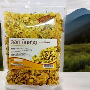 Puede incluir: Una bolsa de pl&aacute;stico transparente llena de flores de crisantemo amarillas secas. La bolsa tiene una etiqueta blanca y amarilla con texto en un idioma extranjero. El fondo muestra una cordillera y follaje verde.