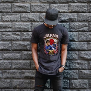 May include: A charcoal gray short-sleeve t-shirt featuring a graphic print with the text JAPAN and FIFA 2026. The design includes two soccer players in blue jerseys, the number 20, and a red circle representing the Japanese flag on a dark background.