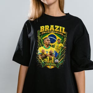 May include: A black t-shirt featuring a graphic print of a soccer player in a yellow Brazil jersey. The design includes the text BRAZIL, the number 11, and FIFA 2026, set against a background of the Brazilian flag, green lightning, and a soccer ball.